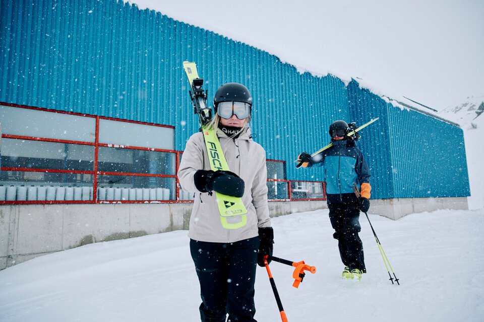 © Fischer Sports GmbH Deux skieurs avec des skis devant un bâtiment bleu dans la neige. | © Fischer Sports GmbH