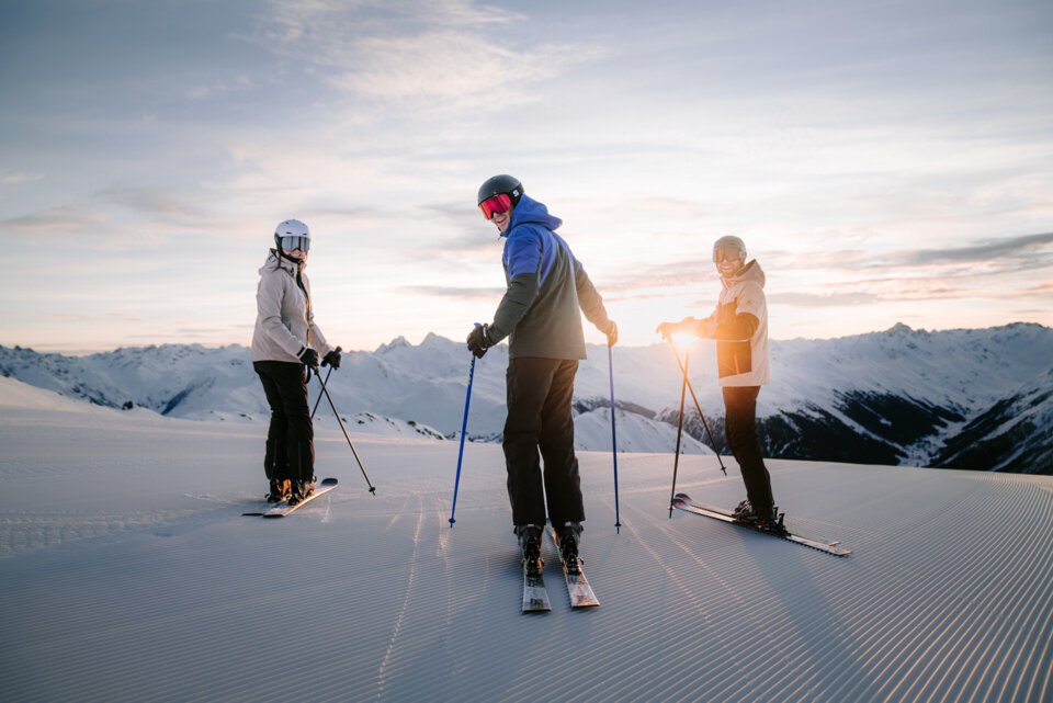 INTERSPORT Rent Salomon Skiing Perfect Ski Day Sunrise Three skiers on a ski slope with snow-covered mountains in the background at sunset.