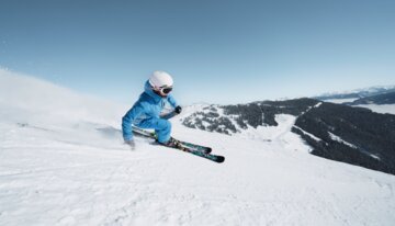 © INTERSPORT International Corporation GmbH Skiër in blauwe jas skiet een besneeuwde helling af. | © INTERSPORT International Corporation GmbH